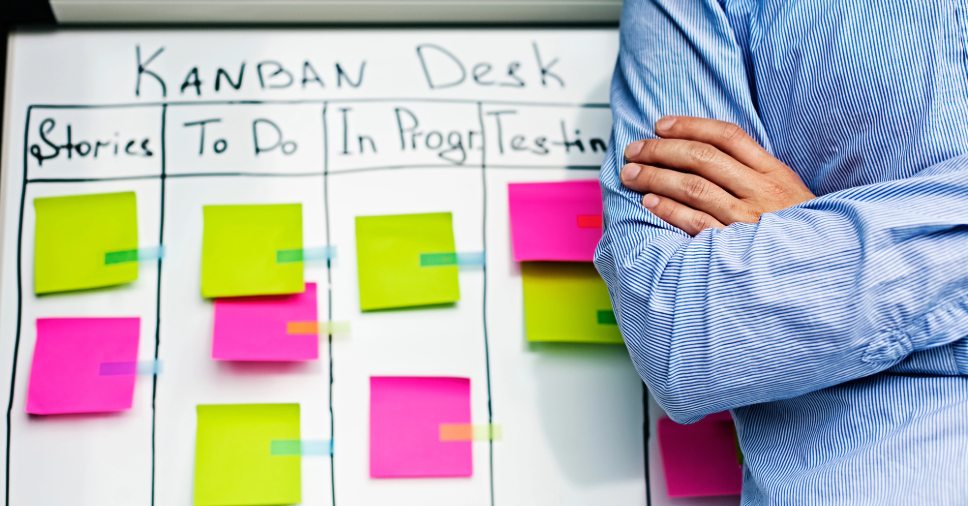 Profissional de braços cruzados ao lado de quadro com post-its de kanban para oficinas.