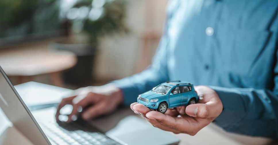 Homem utiliza laptop enquanto segura miniatura de carro azul, simulando uma avaliação virtual.