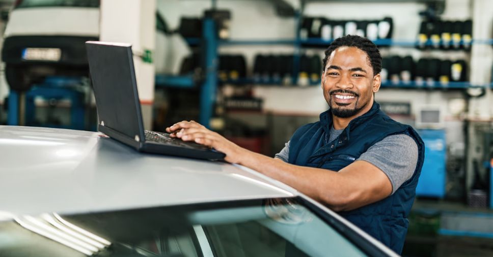 Mecânico sorri com notebook sobre o carro para enviar um lembrete de revisão automotiva.